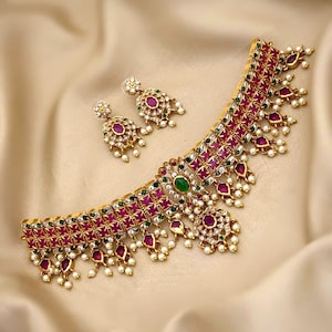 May include: A gold-toned choker necklace and matching stud earrings set featuring square-cut red stones, small green accents, clear crystals, and dangling white pearl-like beads. The text A HOUSE OF UNIQUE DESIGN OF JEWELLERY is at the bottom.