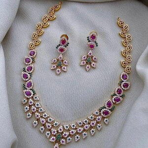 Può includere: Un set composto da collana e orecchini pendenti color oro con pietre rosa e verdi a goccia, circondate da piccoli cristalli bianchi. La collana presenta un motivo a foglie lungo la catena, che si riflette nel design degli orecchini.