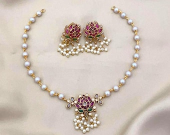 May include: A gold-toned necklace and matching earrings set. The necklace and earrings feature a floral design with red and green accents, surrounded by small white pearls. The jewelry set is displayed on a cream-colored surface.