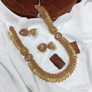 Op de afbeelding: Een goudkleurige ketting en bijpassende oorbellen set. De ketting heeft muntvormige hangers en decoratieve elementen met rode, groene en witte accenten. De oorbellen hebben een vergelijkbaar ontwerp met parelachtige bedels. De sieraden worden getoond op een witte doek.