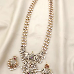 May include: An ornate gold-toned necklace and earring set. The necklace features multiple strands of small, white pearls and a large pendant with intricate detailing, including clear and red gemstones. Matching earrings are also included.