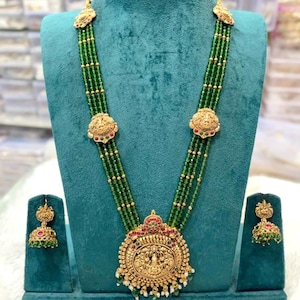 Puede incluir: Un conjunto de collar y pendientes dorados con hilos de cuentas verdes. El collar presenta un colgante grande y ornamentado con una figura central y elementos decorativos. Los pendientes reflejan el diseño del collar, expuestos sobre un soporte de terciopelo verde azulado.