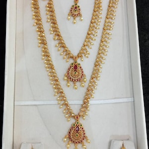 May include: A gold-toned jewelry set featuring two long necklaces, a pair of earrings, and a maang tikka. Each piece is adorned with small white beads and inlaid with red and green stones in a teardrop-shaped pendant design with intricate metalwork.