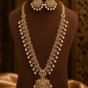 May include: A gold-toned necklace and earring set featuring intricate metalwork, embedded with small white stones, red and green accents, and dangling white pearls. The long necklace includes a large, ornate central pendant with matching drop earrings.