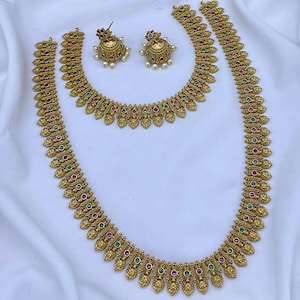 May include: A gold-toned jewelry set consisting of a long necklace, a shorter necklace, and a pair of jhumka earrings. Each piece features intricate metalwork with alternating round red and green stones and small dangling white beads.