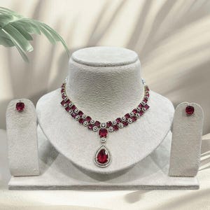 Può includere: Un set di gioielli composto da una collana e orecchini a lobo abbinati. La collana presenta una doppia fila di pietre rosse quadrate circondate da piccoli cristalli bianchi, con un grande ciondolo rosso a forma di goccia al centro.