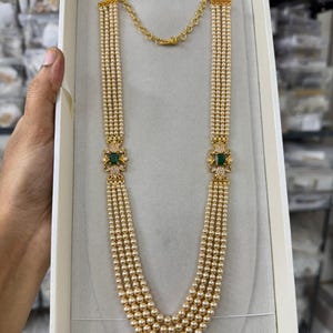 May include: A multi-strand necklace featuring four rows of small cream-colored pearls. Two gold-toned metal connectors are positioned on each side, each set with a rectangular green stone surrounded by small clear crystals and a gold chain extender.