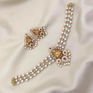 Può includere: Un set di gioielli color oro composto da una collana e orecchini abbinati. La collana ha tre fili di perle bianche e un pendente centrale con una figura dettagliata. Gli orecchini hanno un design simile con accenti rossi.