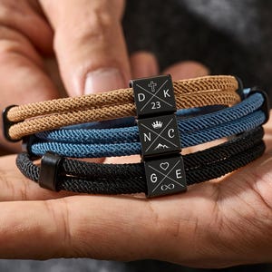 Op de afbeelding: Drie gevlochten touwarmbanden in bruin, blauw en zwart, elk met een zwarte vierkante bedel. De bedels hebben symbolen, initialen en het nummer 23. De armbanden worden in een hand gehouden.
