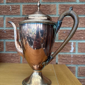 May include: A vintage copper and silver-plated teapot with a wooden handle. The teapot features a curved spout, a lid with a small finial, and a pedestal base. The polished surface reflects the surroundings. The handle is a dark brown colour.