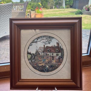 May include: A framed print depicting a house under construction, set within a circular design. The print is in a brown wooden frame and displayed on a clear acrylic stand. The artwork shows a rural scene with workers and vehicles. The frame is approximately 25 cm square.