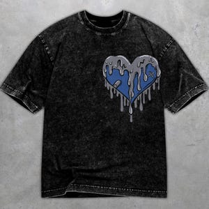 May include: Black acid wash t-shirt featuring a blue heart design with a gray, dripping effect. The heart has a cross and a small gray shape. The shirt has a crew neck and short sleeves.
