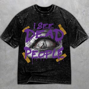 Könnte beinhalten: Schwarzes Acid-Wash-T-Shirt mit einer Grafik eines Auges und den Worten "I SEE DEAD PEOPLE" in Lila. Die Augengrafik zeigt eine Hand, die sich ausstreckt. Lila Farbe tropft vom Text. Gelbes Klebeband akzentuiert das Design.