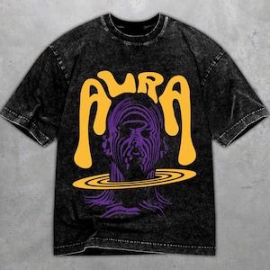 Könnte beinhalten: Schwarzes T-Shirt mit Säurewaschung und Grafikdesign. Das Design zeigt das Wort "AURA" in Gelb über einer lila Illustration eines Gesichts mit einem Ring um den Hals. Das T-Shirt hat kurze Ärmel und einen Rundhalsausschnitt.