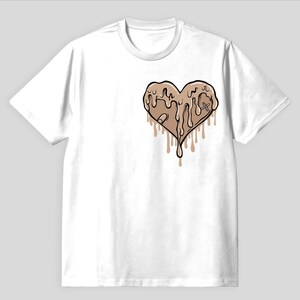 Könnte beinhalten: Weißes T-Shirt mit einer Grafik eines braunen Herzens mit tropfenden Details. Das Herz hat ein Pflaster und ein Kreuz-Design. Das T-Shirt hat einen Rundhalsausschnitt und kurze Ärmel. Das Design befindet sich auf der linken Brustseite.
