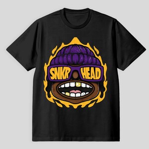 Könnte beinhalten: Schwarzes T-Shirt mit einem Cartoon-Gesicht. Das Gesicht hat eine lila Mütze, gelbe Sonnenbrille mit dem Text "SNKR HEAD" und ein gelbes Flammendesign. Das Design hat einen braunen Hautton und einen offenen Mund.