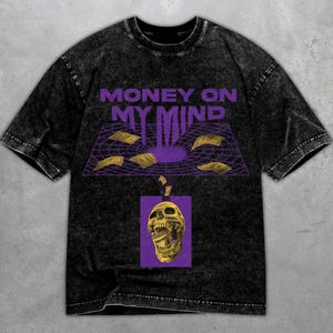 Könnte beinhalten: Schwarzes Grafik-T-Shirt mit dem lila Schriftzug "MONEY ON MY MIND". Das Design zeigt ein lila Gitter mit fallendem Geld und einen goldenen Totenkopf auf lila Hintergrund. Das Shirt hat einen Vintage-Look.