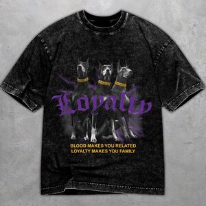Könnte beinhalten: Schwarzes T-Shirt mit einem Grafikdesign von drei Dobermann Pinschern mit goldenen Halsbändern und dem Wort "Loyalität" in lila Schrift. Der Text "Blut verbindet, Loyalität macht zur Familie" ist ebenfalls vorhanden.