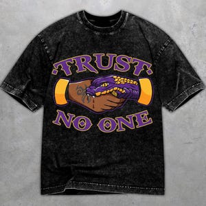 Könnte beinhalten: Schwarzes T-Shirt mit Säurewaschung und Grafikdesign. Das Design zeigt einen Händedruck zwischen einer braunen Hand und einem lila Schlangenkopf mit den Worten "TRUST NO ONE" in lila und goldener Schrift.