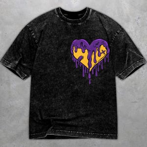 Könnte beinhalten: Schwarzes Acid-Wash-T-Shirt mit einem gelben Herzdesign und lila tropfenden Akzenten. Das Herz hat ein kleines Kreuz auf der rechten Seite. Das T-Shirt hat kurze Ärmel und einen Rundhalsausschnitt.