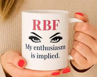 Può includere: Tazza in ceramica bianca con la scritta "RBF" in rosa, occhi stilizzati e la frase "My enthusiasm is implied." La tazza è tenuta da una persona con lo smalto rosso.