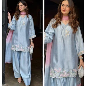 May include: A light blue silk salwar kameez set with intricate embroidery. The set includes a long-sleeved tunic, loose-fitting pants, and a matching scarf. The tunic features floral embroidery around the neckline and cuffs, and the pants have embroidered detailing at the hem. The text "RiddhiSiddhiTextile" is at the top.