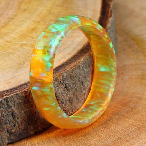May include: A translucent orange ring with green and blue swirls. The ring's iridescent colours are set against a natural wood background. This striking piece of jewellery is a unique accessory, perfect for adding a pop of colour.