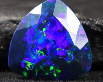 May include: A triangular black opal gemstone with a polished, faceted surface. The opal exhibits vibrant internal colours of blue, purple, and green, creating a dynamic play of light. The gemstone is set against a dark background.