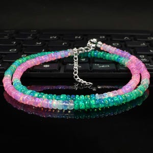 Puede incluir: Un collar de cuentas con pequeñas cuentas de ópalo redondas e iridiscentes en tonos rosa, verde y azul. El collar incluye una cadena extensora de metal plateado con un cierre de mosquetón para un ajuste regulable y un cierre seguro.