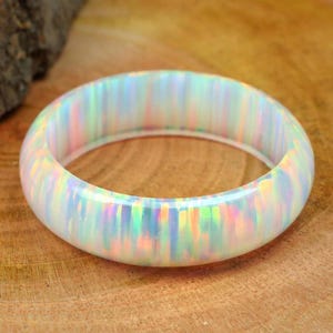 May include: A white opal bangle bracelet with iridescent color flashes. The smooth, rounded bracelet is set against a wooden surface. The opal displays a range of colors, including pink, green, and blue, creating a shimmering effect.