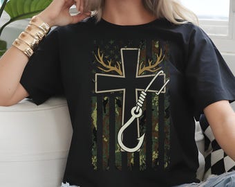 May include: Black t-shirt featuring a cross design with deer antlers and a fishing hook. The cross is overlaid on a camouflage pattern resembling a flag. The shirt is a casual wear item.