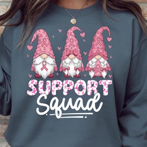 May include: A teal sweatshirt featuring three gnomes with pink hats and white beards. The gnomes are decorated with pink ribbons and hearts. The words "SUPPORT Squad" are printed in white and pink.