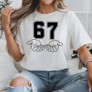 May include: White t-shirt featuring the number "67" in a black, block-style font with a white outline. Below the numbers are two open hands in a line drawing. The shirt is worn by a person wearing blue jeans.