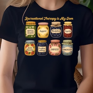 以下が含まれることがあります： 「Recreational Therapy is My Jam」の文字が入った濃いネイビーブルーのTシャツ。シャツには、それぞれ「Mental Health」や「Empathy」など、メンタルヘルスに関する様々な側面がラベル付けされた、8つのジャム瓶のイラストが描かれています。