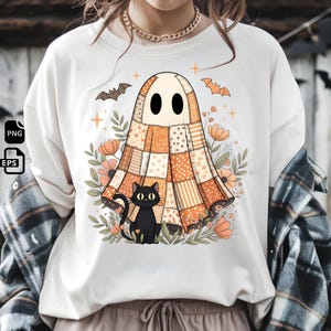 May include: Cream-colored t-shirt featuring a Halloween-themed graphic. The design includes a patchwork ghost, a black cat, bats, flowers, and stars. The ghost is in shades of orange, cream, and brown. The cat is black, and the flowers are peach.
