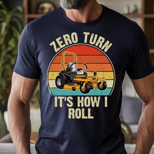 May include: Navy blue t-shirt featuring a graphic of a yellow zero-turn lawn mower set against a sunset design. The text reads "ZERO TURN" above the mower and "IT'S HOW I ROLL" below. The shirt is a casual wear item.