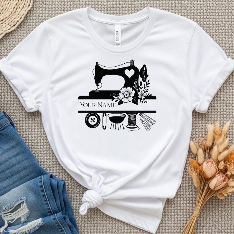 Pode incluir: Camiseta branca com um gr&aacute;fico preto de uma m&aacute;quina de costura, um cora&ccedil;&atilde;o, flores e utens&iacute;lios de costura. O texto "YOUR NAME" &eacute; exibido abaixo da m&aacute;quina de costura. Um bot&atilde;o, uma agulha, um carretel de linha e uma fita m&eacute;trica tamb&eacute;m est&atilde;o inclu&iacute;dos.