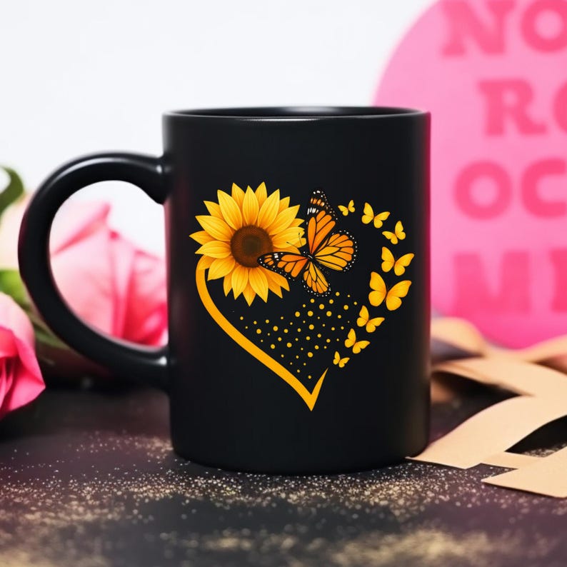 May include: Black ceramic mug featuring a yellow sunflower and orange monarch butterflies arranged in a heart shape. The mug has a black handle and a glossy finish. The design includes a dotted trail of butterflies.