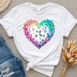 May include: White t-shirt with a heart-shaped design made of colourful butterflies and flowers. The butterflies are in shades of pink, purple, blue, and green, forming a rainbow. The shirt is made of a soft material and has short sleeves.