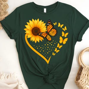 May include: Dark green t-shirt featuring a yellow sunflower and monarch butterfly design. The design includes a yellow heart shape, small yellow butterflies, and yellow dots. The shirt is knotted at the waist.