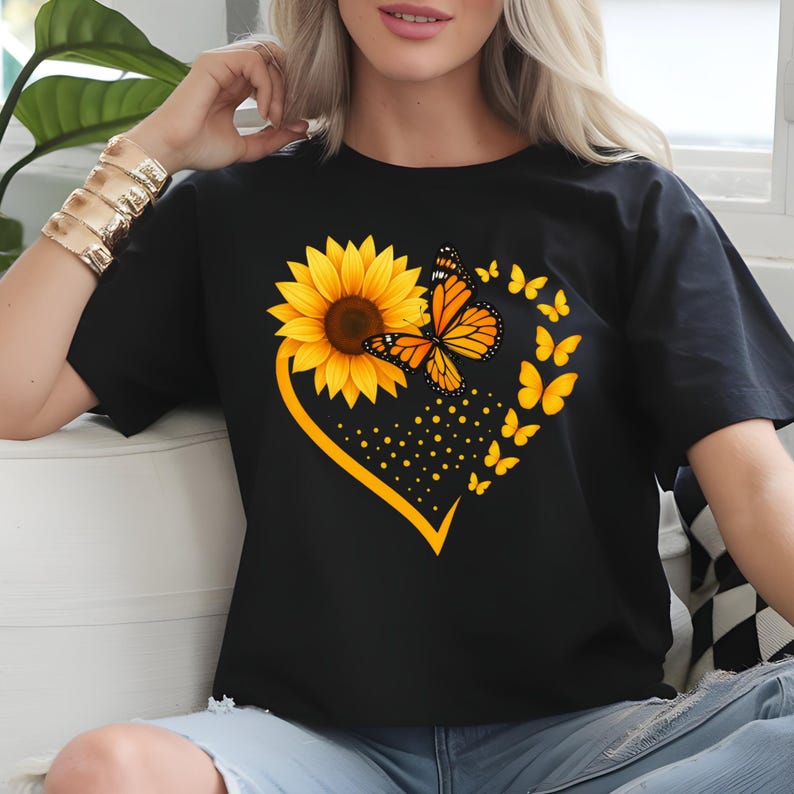 May include: Black t-shirt featuring a heart-shaped design with a yellow sunflower and orange butterfly. The design includes a cluster of yellow butterflies and yellow dots. The shirt is a casual wear item.