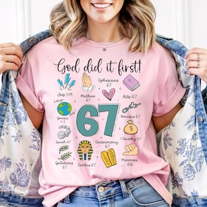 May include: A light pink t-shirt with the text "God did it first!" and the number "67" in teal. The shirt features various illustrations and Bible verses. The shirt is made of a soft material.