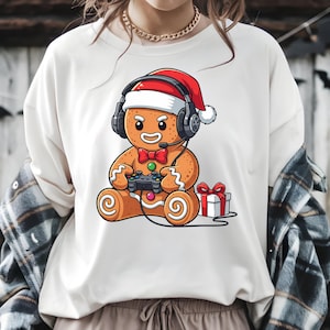 May include: A cream-colored sweatshirt featuring a cartoon gingerbread man wearing a Santa hat and headphones, holding a video game controller. A red bow tie and a wrapped gift box are also visible. The design is festive and playful.