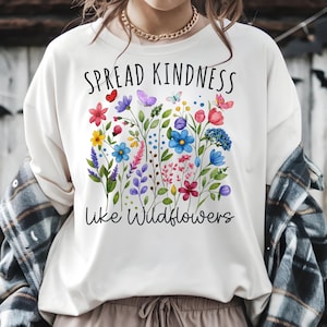 May include: A cream-coloured sweatshirt featuring a floral design and the text "SPREAD KINDNESS like Wildflowers." The graphic includes colourful flowers and butterflies in shades of red, blue, purple, and yellow.
