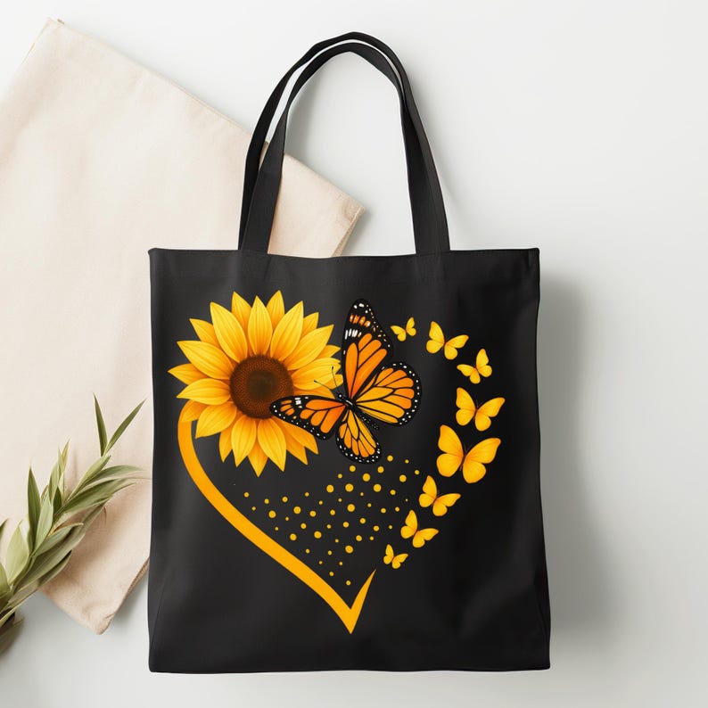 May include: Black tote bag featuring a sunflower and monarch butterfly design. The sunflower is yellow with a brown center, and the butterfly is orange and black. A yellow heart shape surrounds the sunflower and butterfly, with smaller yellow butterflies trailing.