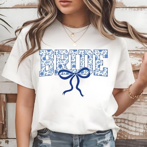 Puede incluir: Camiseta blanca con la palabra "BRIDE" en estampado floral azul. Un lazo azul está centrado debajo de la palabra. La camiseta es de cuello redondo y está hecha de un material suave.