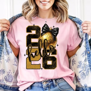 以下が含まれることがあります： 「hello 2026」の文字が黒と金でプリントされた、薄いピンク色のTシャツ。 「hello」という単語の上に、黒いリボンが付いた金のディスコボールが描かれています。 このシャツは半袖のクルーネックです。