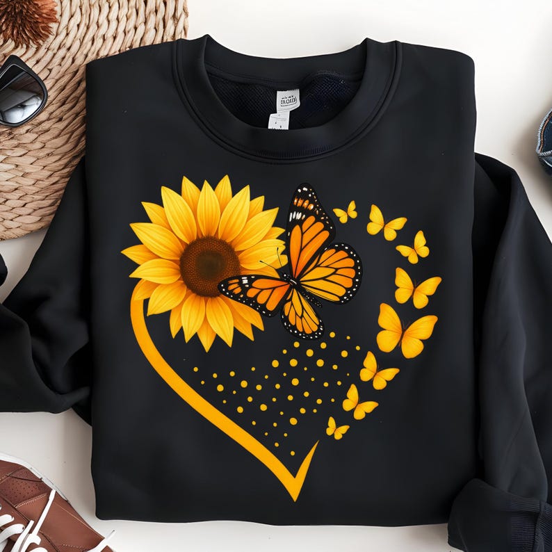 May include: Black sweatshirt featuring a sunflower and monarch butterfly design. The sunflower is yellow and brown, with a monarch butterfly in orange and black. A heart shape is formed with a yellow line and butterflies.