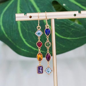 Puede incluir: Pendientes colgantes dorados con una serie de gemas de colores. Los pendientes presentan una variedad de formas, incluyendo cuadrados, diamantes, corazones y óvalos, en tonos de azul, rojo, naranja, verde y morado. Los pendientes se exhiben en un soporte dorado.