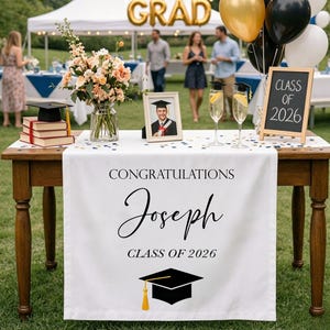 Puede incluir: Una mesa de fiesta de graduación está decorada con un camino de mesa blanco que dice "Felicitaciones Joseph Clase de 2026". Una foto enmarcada, flores, copas de champán y globos decoran la mesa. Una pequeña pizarra dice "Clase de 2026".